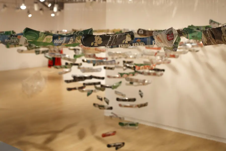 A series of miniature canoe sculptures with beer branding are hanging from the ceiling in an art gallery.