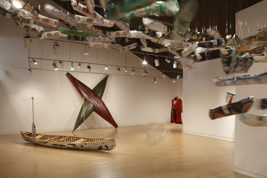 View of an art exhibition with many miniature canoe sculptures hanging from the ceiling. From left to right: mirrored canoe, two intersecting canoes, glass canoe sculpture and hanging coat in the background.