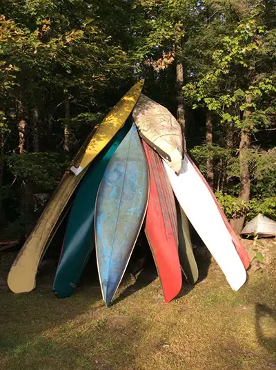 Several canoes are piled up to create a pyramid-shaped structure in front of a forested area.