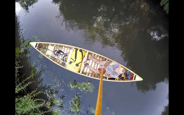 Artist Brad Copping holding a paddle as a camera pole creating a bird’s eye view of him in his canoe in the water.