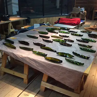 Many miniature green glass canoe sculptures lay on a table with a folded sheet of red fabric in the background.