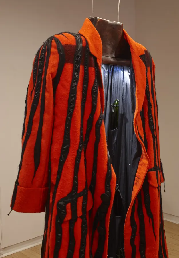 An open red coat with black stripes is hanging from the ceiling with a light inside illuminating the fabric. A small shiny green object is tucked in a pouch inside the coat.