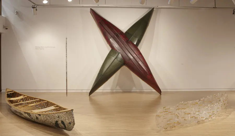 Two canoes, one red and one green, intersect to form an 'X' shape. A canoe with mirror fragments and a glass canoe sculpture lay on the gallery floor on either side.