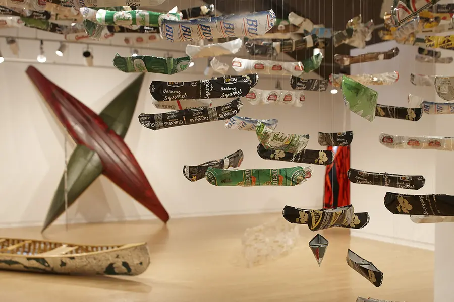 A series of miniature canoe sculptures with beer branding are hanging from the ceiling in an art gallery.