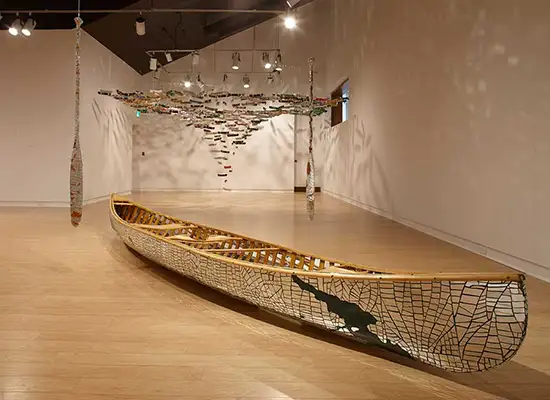 A canoe with mirror fragments sculpture lay on the gallery floor with small canoes hanging in the background.