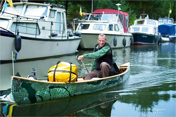 Artist Brad Copping smiling at camera while paddling a canoe with mirror fragments beside a lineup of boats.