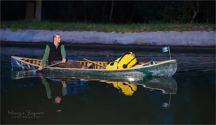 Side view of artist Brad Copping paddling a canoe with mirror fragments at nighttime.