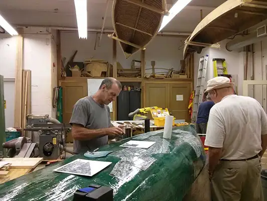 Two people attaching mirror fragments to the underside of a canoe in a workshop.